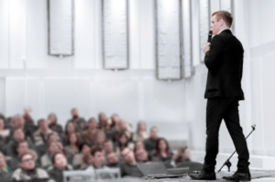 Imagem da notícia Imagem de homem de terno preto, em pé em espécie de palco, falando ao microfone para grande grupo de pessoas sentadas na plateia