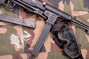 Imagem da Notícia Imagem de arma de airsoft preta ao lado de esferas de plástico espalhadas e óculos de proteção